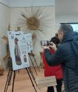 Чоловік у темній куртці фотографує на смартфон виставковий стенд із малюнком жінки в традиційному українському вбранні, встановлений на тринозі в музейній кімнаті з плетеними з лози виробами на задньому плані.