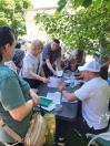 Група людей на вулиці під деревом зібралася навколо столу, де чоловік у білій кепці та інші організатори приймають документи, заповнюють папери та спілкуються з громадянами