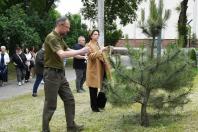 Чоловік у зеленій футболці поло та жінка в бежевому пальті пов'язують символічні стрічки на гілки молодої сосни на галявині парку, тоді як на задньому плані за ними спостерігають інші люди.