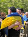 Два українських військовослужбовці у чорних кепках емоційно та міцно обіймають один одного на вулиці, загорнувшись у синьо-жовтий прапор України під час повернення з полону.