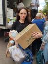 Жінка в чорній футболці стоїть на вулиці біля вантажівки та тримає в руках велику картонну коробку з логотипом «bonya» та пакети з отриманою гуманітарною допомогою, поки на задньому плані волонтери продовжують видачу.