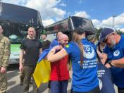 Представники Координаційного штабу у синіх футболках зустрічають звільнених із полону українських військовослужбовців біля великих автобусів, накидаючи їм на плечі державні синьо-жовті прапори.