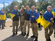 Чотири звільнених з полону українських військовослужбовці у камуфляжних штанах та із синьо-жовтими прапорами на плечах радісно позують для фото, обійнявшись у шеренгу на асфальтованому майданчику на тлі лісу.