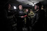 У тьмяно освітленому підземному укритті журналіст у бронежилеті з написом «PRESS» тримає мікрофон із червоним логотипом і бере інтерв'ю у чоловіка в чорній куртці, поки оператор знімає процес, а на задньому плані стоять військовослужбовці.
