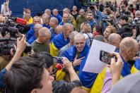 Великий натовп журналістів, фотографів та цивільних осіб із камерами, мікрофонами й телефонами оточив групу звільнених із полону українських військовослужбовців із поголеними головами, які загорнуті в синьо-жовті прапори та щиро посміхаються
