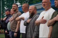 Шеринг чоловіків середнього віку, серед яких бородатий посадовець Олексій Харченко, стоїть на відкритому спортивному майданчику, урочисто приклавши праву руку до серця під час виконання державного гімну України.