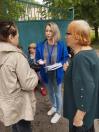На вулиці біля високих зелених воріт світловолоса жінка у синій кофті з бейджем на шиї тримає інформаційні картки та розмовляє з двома жінками, що стоять поруч із нею