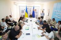 У світлому залі за довгим Т-подібним столом проходить офіційна зустріч або пресконференція численної групи людей за участю представників Луганської ОВА, на тлі банерів «Official Media Center» та державних прапорів.