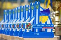 Ряд однакових синьо-білих сувенірних нагород Луганської області, прикрашених малим гербом України, картою регіону у синьо-жовтих кольорах, офіційним гербом області, орнаментом та цифрами «1938», вишикувані на дерев'яній підставці