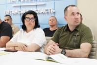 На тлі блакитної стіни з написом «Луганська область» за білим столом сидять люди, серед яких на передньому плані виділяються жінка в окулярах та білій футболці й чоловік у зеленій футболці-поло, які уважно слухають або дивляться вбік.