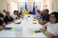 У світлому кабінеті за довгим білим столом сидить група людей під час наради чи брифінгу, на чолі якого в торці зали під прапорами та офіційними банерами перебуває керівник.