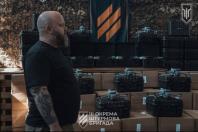 На зображенні бородатий чоловік із татуюванням на руці стоїть у профіль на тлі численних картонних коробок із пластиковими кейсами та квадрокоптерами, а також прапора та емблеми з написом «ІІІ ОКРЕМА ШТУРМОВА БРИГАДА».