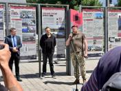 На світлині просто неба чоловік у камуфляжній формі та бордовому береті виступає з мікрофоном біля стійки перед великими інформаційними стендами виставки, а ліворуч від нього слухають двоє чоловіків — один у піджаку та окулярах, інший у чорній кофті.