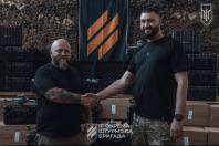 На світлині двоє бородатих чоловіків у чорних футболках тиснуть один одному руки на тлі картонних коробок із кейсами для обладнання, прапора та емблеми з написом «ІІІ ОКРЕМА ШТУРМОВА БРИГАДА».