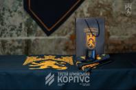 На фото зображено стіл, накритий темною скатертиною з помаранчевим логотипом у вигляді лева та фортеці, на якій стоїть подарунковий пакет, відкрита коробочка з відзнакою та складений прапор із такою ж символікою.