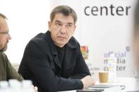 На фото зображено чоловіка середнього віку з короткою стрижкою у темній куртці, який сидить за столом на тлі світлого банера з написом «official media center».
