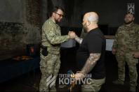 На фото зображено чоловіка у військовій формі камуфляжу «мультикам», який посміхається та тисне руку лисому чоловікові у чорній футболці з татуюваннями на руках, у темному приміщенні з логотипом «Третій армійський корпус».