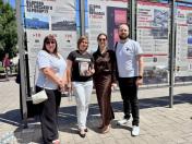 На світлині просто неба четверо людей — три жінки та чоловік — стоять у ряд і посміхаються на тлі великих інформаційних стендів про оборону Луганського аеропорту, при цьому одна з жінок по центру тримає в руках книгу