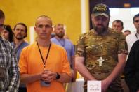 Двоє чоловіків — один у яскраво-помаранчевій сорочці зі складеними разом долонями, а інший у камуфляжній футболці, бейсболці та з великим хрестом на шиї — стоять із серйозними виразами облич серед інших людей у залі.