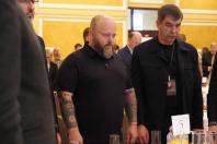 Урочисто вбрані учасники офіційного заходу стоять біля круглого сервірованого столу з номером п'ять, схиливши або опустивши голови в момент тиші чи молитви, де на передньому плані виділяються двоє чоловіків у чорному одязі, один із яких має татуювання на руках та густу бороду.