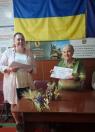 У кімнаті на тлі українського прапора та інформаційних плакатів літня жінка в традиційній вишиванці сидить за столом і тримає сертифікат, тоді як поруч стоїть усміхнена молода жінка з аркушем паперу, а на самому столі лежать декоративні колоски.