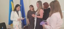 П'ять жінок спілкуються в кабінеті на тлі прапорів України та Європейського Союзу, тримаючи в руках документи та брошури під час офіційної зустрічі.