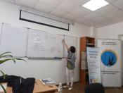 У світлій навчальній аудиторії жінка стоїть спиною до камери та пише маркером схеми на великій білій дошці, поруч із якою розміщено банери громадської організації «Десяте квітня»