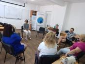 У світлій аудиторії з білою дошкою та банерами громадської організації «Десяте квітня» група жінок різного віку сидить на стільцях у колі під час групового заняття чи тренінгу.