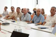 На зображенні група людей, переважно чоловіків середнього та старшого віку, сидить у світлій аудиторії за довгим білим столом із блокнотами, смартфонами та пляшками води, уважно слухаючи спікера під час наради чи семінару
