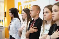 Група молодих людей стоїть у профіль в один ряд, приклавши праві руки до серця під час урочистого моменту чи виконання гімну.