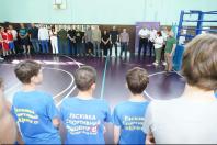 Група юних спортсменів у синіх футболках стоїть спиною до камери та спостерігає за офіційною лінією дорослих і суддів, які вишикувалися вздовж стіни спортивного залу біля боксерського рингу.