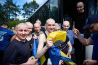 Група усміхнених та виснажених українських військовослужбовців із поголеними головами виходить з автобуса під час обміну полоненими, тримаючи в руках синьо-жовтий державний прапор.