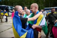 Двоє звільнених з полону українських захисників із поголеними головами, загорнуті у державні прапори України, радісно обіймаються та посміхаються один одному на тлі автобусів та інших військових.