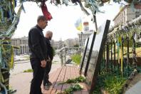 На світлині зображено двох чоловіків у темному одязі, які схилили голови у хвилині мовчання на Алеї Героїв Небесної Сотні у Києві перед гранітними стелами з портретами загиблих, біля підніжжя яких лежать білі троянди.