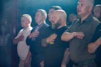 На світлині зображено ряд чоловіків, серед яких є цивільні, службовці та чоловік у вишиванці, котрі зосереджено стоять пліч-о-пліч у задимленому приміщенні та тримають праві руки на серці під час урочистого моменту.