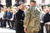 На світлині зображено Президента України Володимира Зеленського в чорному одязі, який тисне руку військовослужбовцю у камуфляжній формі та береті під час урочистої церемонії нагородження на тлі розмитих фігур гостей.