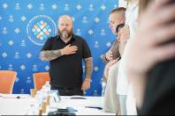 Чоловік із татуюваннями на руках та інші присутні стоять у приміщенні, приклавши праві руки до серця, на тлі синього брендованого банера Конгресу місцевих та регіональних влад.