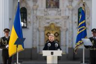 На світлині зображено Президента України Володимира Зеленського, який виступає за білою трибуною з гербом перед величним церковним фасадом, у супроводі двох почесних вартових у парадній формі, котрі тримають державний прапор та штандарт Президента.