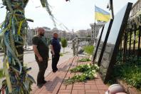 На світлині зображено двох чоловіків — одного в зеленій футболці, а іншого в синій формі ДСНС України, які стоять у хвилині мовчання на Алеї Героїв Небесної Сотні в Києві перед нахиленими чорними гранітними плитами, біля підніжжя яких лежать свіжі білі троянди.
