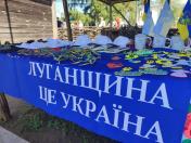Стіл, накритий яскраво-синьою скатертиною з великим білим написом «ЛУГАНЩИНА ЦЕ УКРАЇНА», заставлений патріотичною сувенірною продукцією, прапорцями, чашками та білими кепками під час заходу просто неба.
