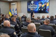 У залі засідань з прапорами поліцейський виступає перед сидячою спиною до кадру аудиторією колег у формі на тлі великого екрана з привітанням «З Днем Національної поліції України!».