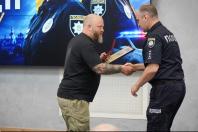 У залі засідань офіцер поліції у формі тисне руку та вручає нагороду у футлярі та грамоту в рамці бородатому чоловікові в чорній футболці з татуюваннями на руці на тлі великого екрана з тематичним зображенням.