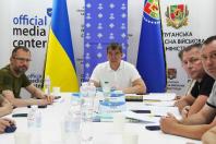 На фотографії зображено робочу зустріч чотирьох чоловіків за білим столом у медіацентрі Луганської обласної військової адміністрації, де по центру сидить керівник Артем Лисогор у білій футболці на тлі державних прапорів, банерів із логотипами, документів та пляшок із водою.