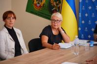 На фотографії зображено двох жінок за дерев'яним столом під час засідання, де одна з них уважно слухає в білому жакеті, а інша, в окулярах та чорній сукні, тримає ручку біля блокнота на тлі державного прапора України, синього банера Конгресу та зеленого військового прапора 3-го прикордонного загону.