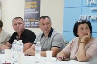На фотографії зображено трьох учасників офіційного засідання — двох чоловіків та жінку, які сидять за білим столом із пляшками води й паперовими склянками та уважно дивляться в один бік на тлі блакитної стіни з написом «Луган...» і плаката з подякою ветеранам.