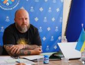 На фотографії зображено міцного бородатого чоловіка з татуюваннями на обох руках, який сидить за робочим столом із ноутбуком, документами та маленьким прапором України на тлі великого синього банера з логотипами Палати місцевих влад.