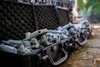 На передньому плані великим планом показано сірий квадрокоптер із потужним об'єктивом камери, який лежить у відкритому чорному захисному кейсі з рельєфним поролоном на кришці, а далі в ряд на землі вишикувані інші аналогічні розпаковані дрони в кейсах на тлі розмитої зелені.
