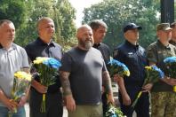 Група чоловіків у цивільному одязі, поліцейській та військовій формі стоїть у шерензі, зосереджено тримаючи в руках патріотичні синьо-жовті букети квітів під час офіційного заходу вшанування пам'яті на тлі зелених дерев.