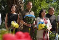 Чотири людини — двоє чоловіків та дві жінки, одна з яких одягнена у вишиванку — стоять поруч із серйозними виразами облич, тримаючи в руках патріотичні синьо-жовті букети квітів під час офіційного заходу вшанування пам'яті на тлі дерев.