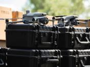 На зображенні представлено два сірі квадрокоптери з розкладеними пропелерами та камерами, які стоять на великих чорних захисних пластикових кейсах, складених у два ряди на тлі картонних коробок та розмитої зелені.